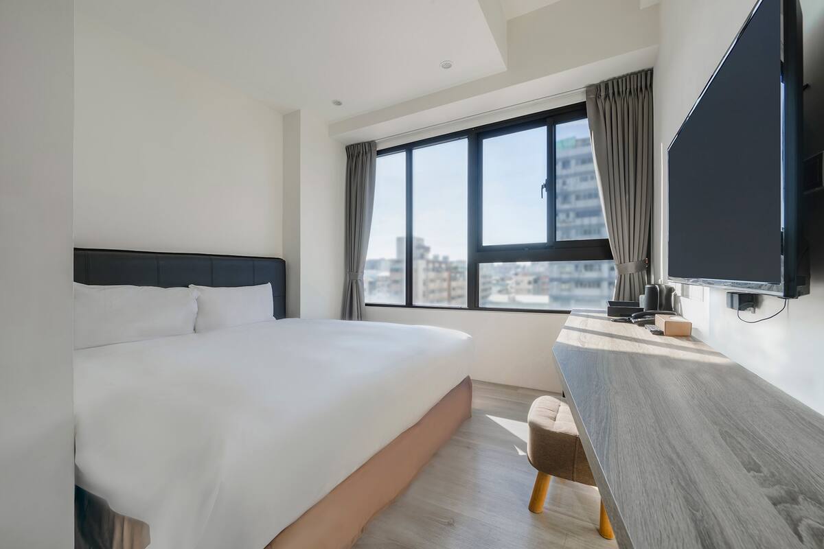 A modern hotel room with a large bed, a desk with a flat-screen TV, and a view of the city through large windows.