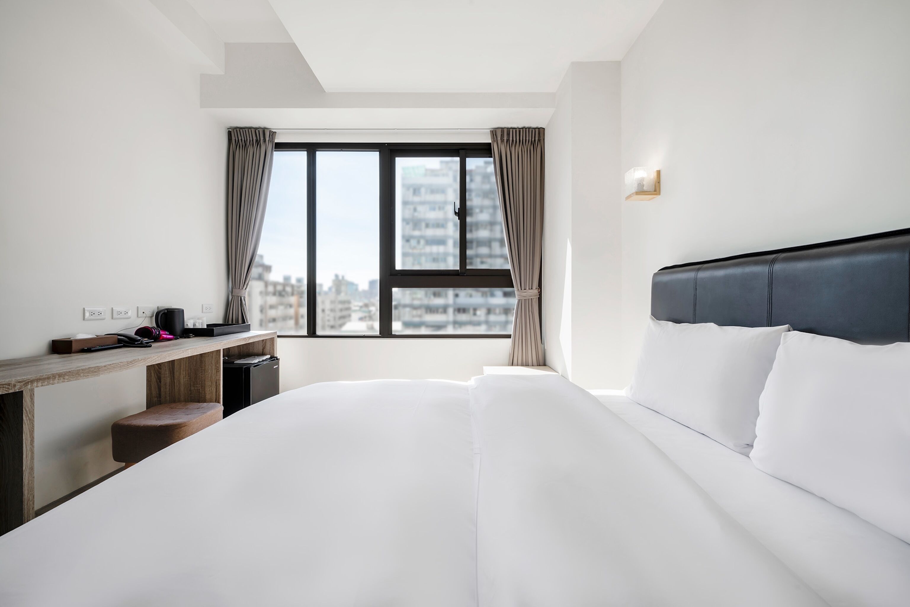 Quarto de hotel moderno com uma cama grande, uma escrivaninha de madeira e vista para a cidade através de uma janela ampla.