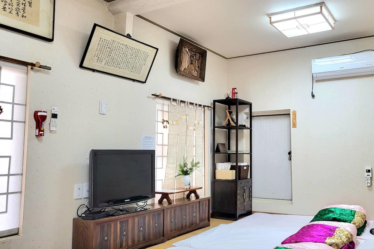 A bedroom with a bed, a television on a wooden cabinet, a bookshelf, a window with curtains, and an air conditioner on the wall.