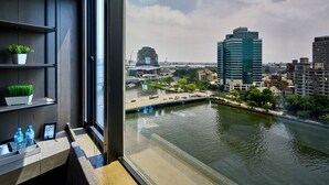 Habitación de hotel moderna con un amplio ventanal que da a un río y al paisaje urbano, incluyendo un edificio distintivo y un puente.