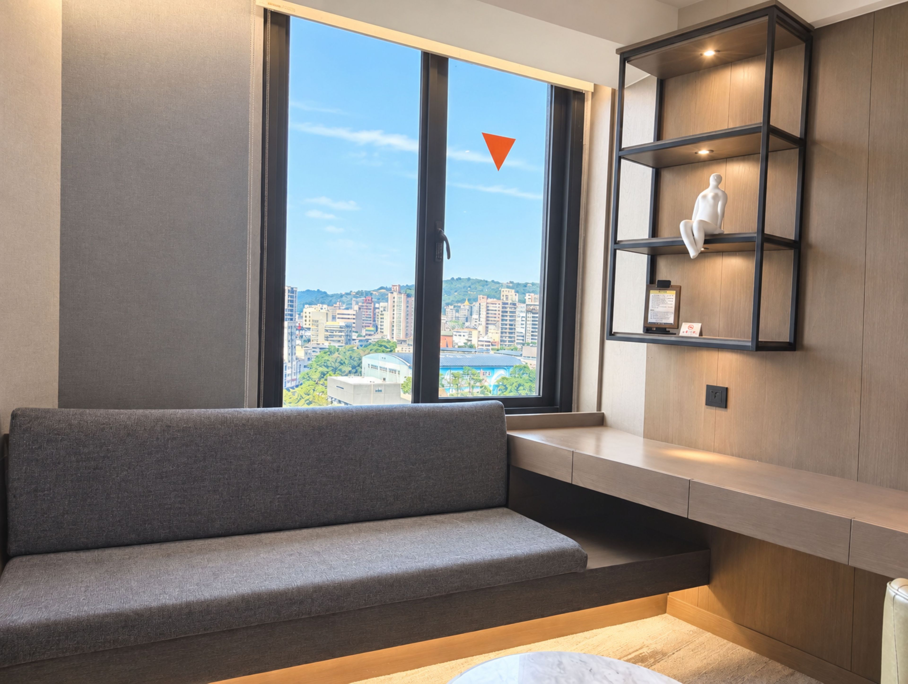 Una habitación moderna con un sofá gris, un estante de madera con un maniquí y un ventanal con vistas a un paisaje urbano.