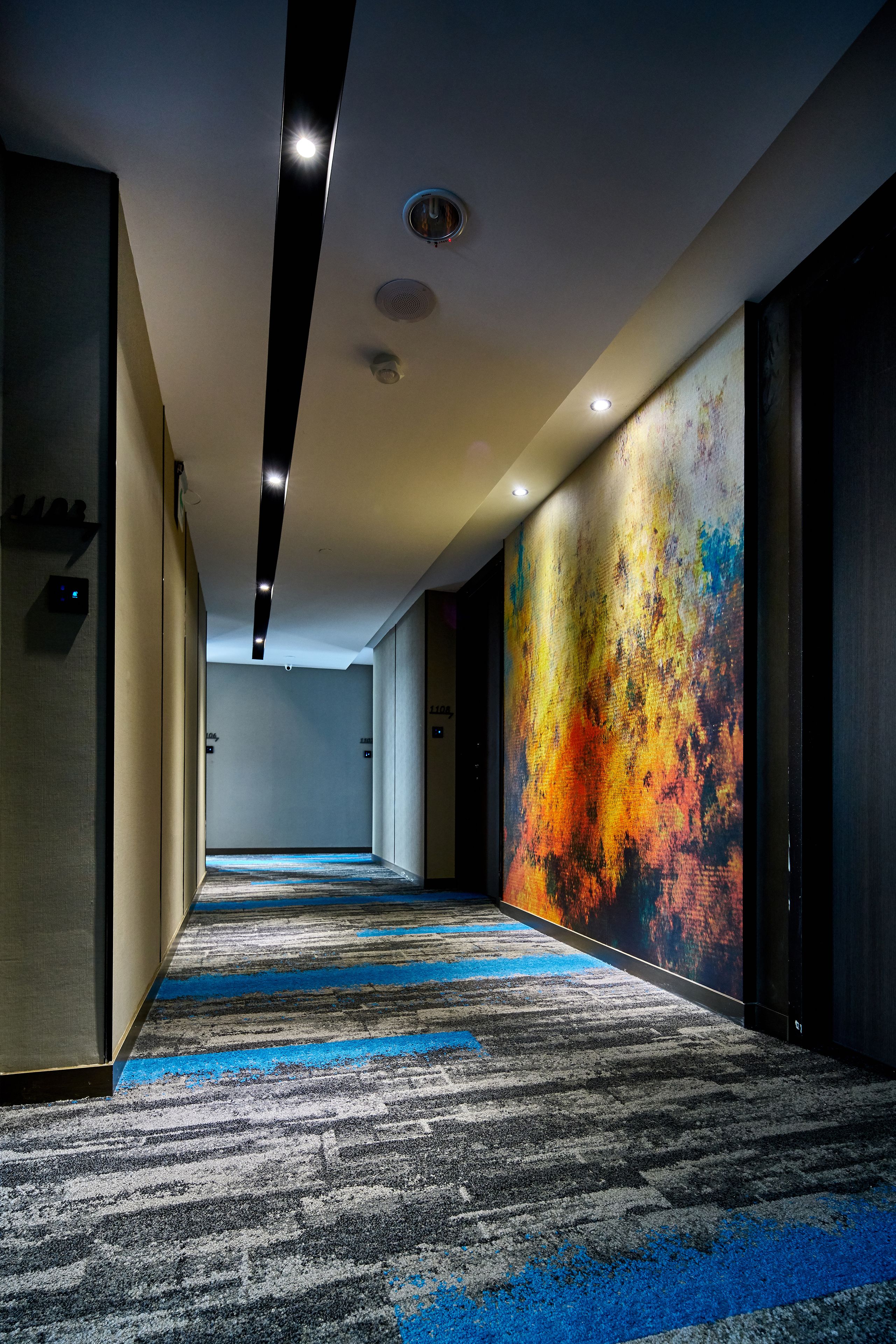 Un couloir d’hôtel avec un tapis à motifs, une fresque murale colorée et un design de plafond moderne.