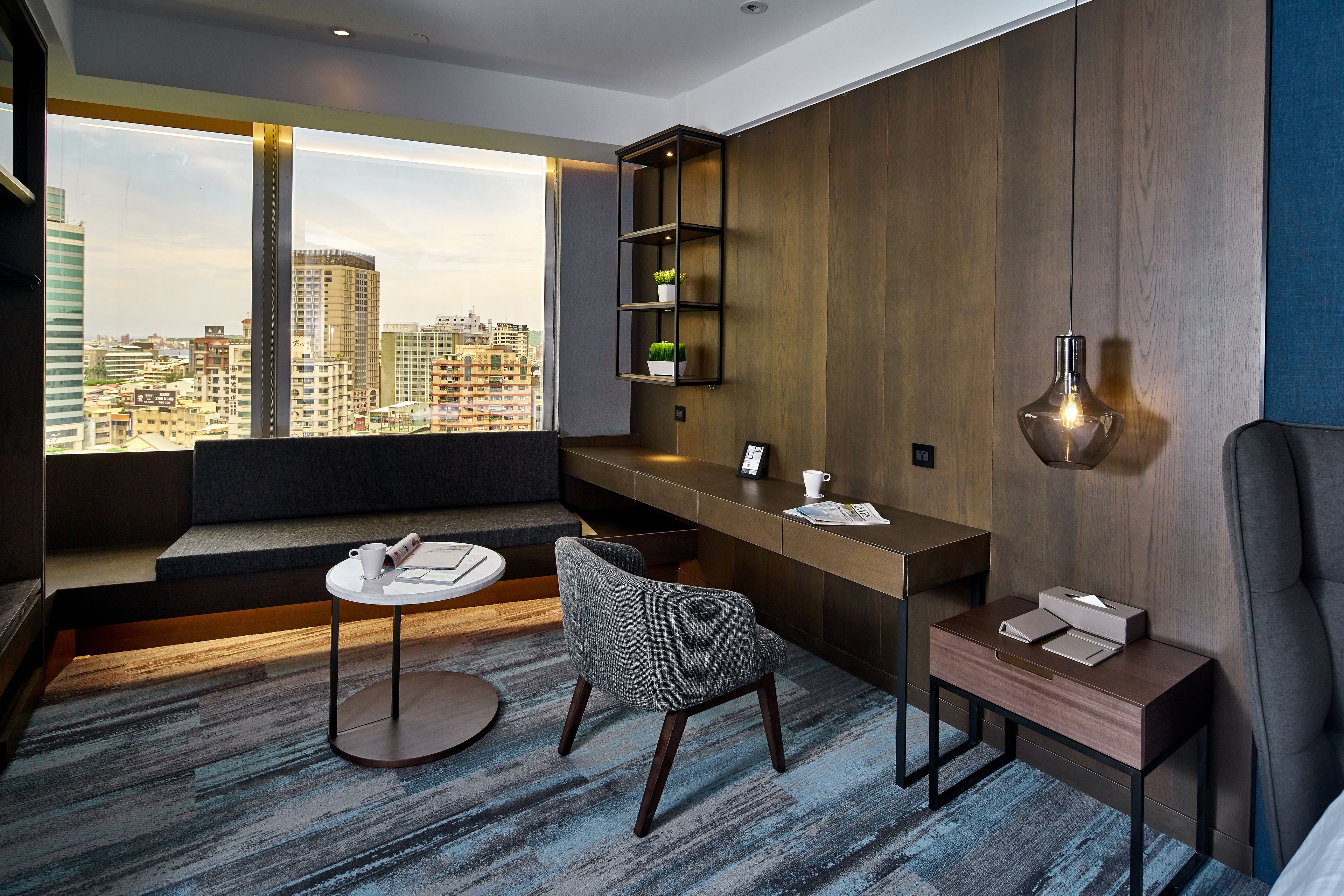 Ein modernes Hotelzimmer mit Blick auf die Stadt, einem Schreibtisch, einem bequemen Sessel, einem kleinen Tisch und einem Nachttisch mit Lampe.
