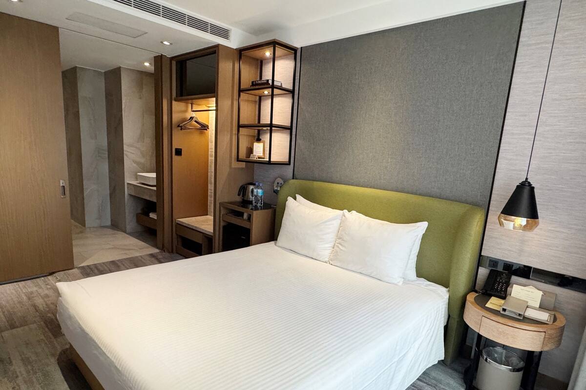 Habitación de hotel con una cama grande, cabecera verde, mesita de noche con lámpara, una pequeña mesa con hervidor y un armario.