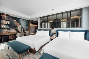 Una camera d'albergo con due letti, una grande finestra, un quadro appeso e una scrivania con una lampada.