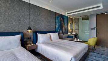Une chambre d’hôtel avec deux lits, une table de chevet, une chaise, un bureau et une salle de bain visible à travers une porte en verre.