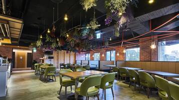 A modern restaurant interior with green chairs, wooden tables, hanging plants, and brick walls.
