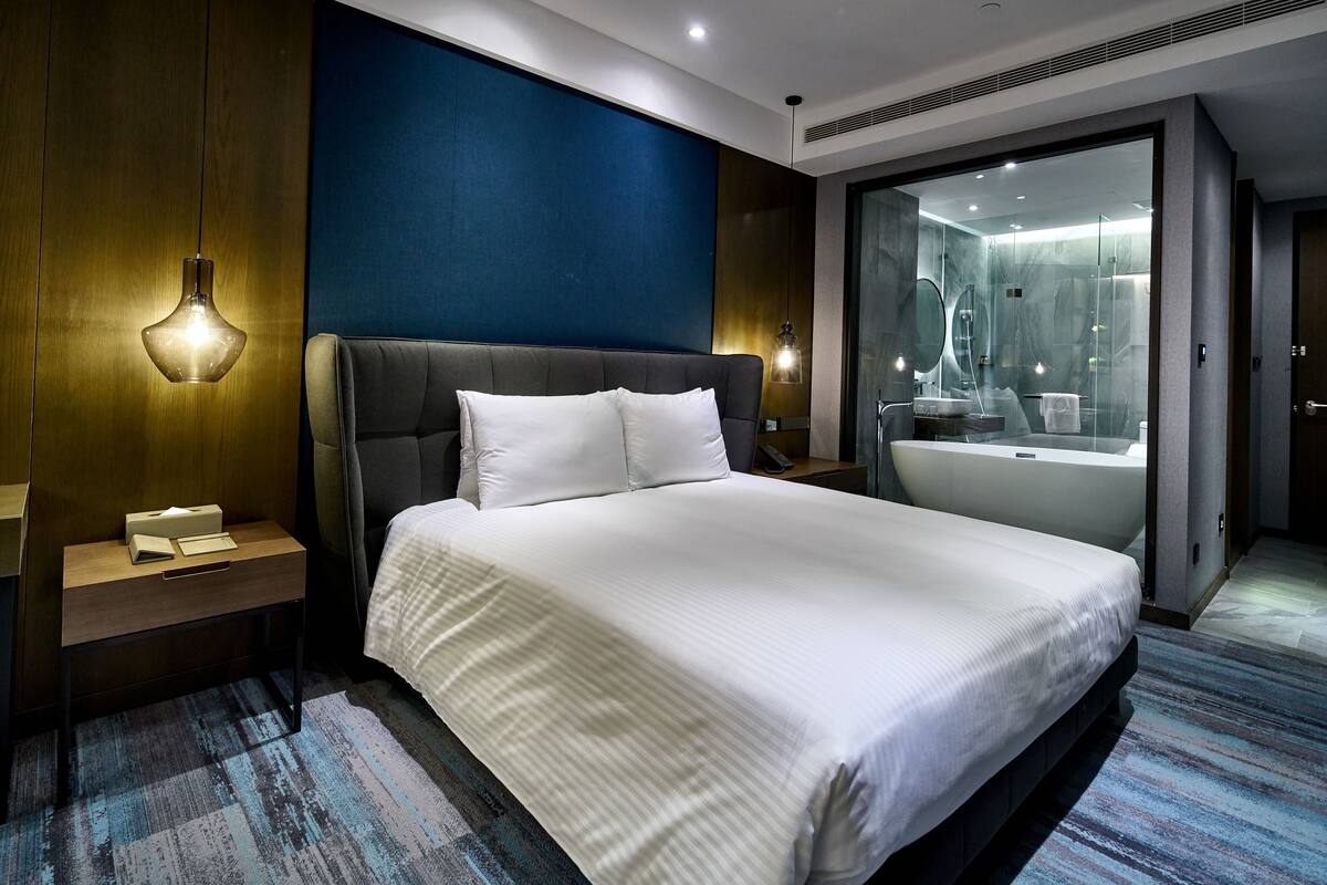Una habitación de hotel moderna con una cama grande, una mesita de noche, una lámpara de pared y un baño con bañera y mampara de vidrio.