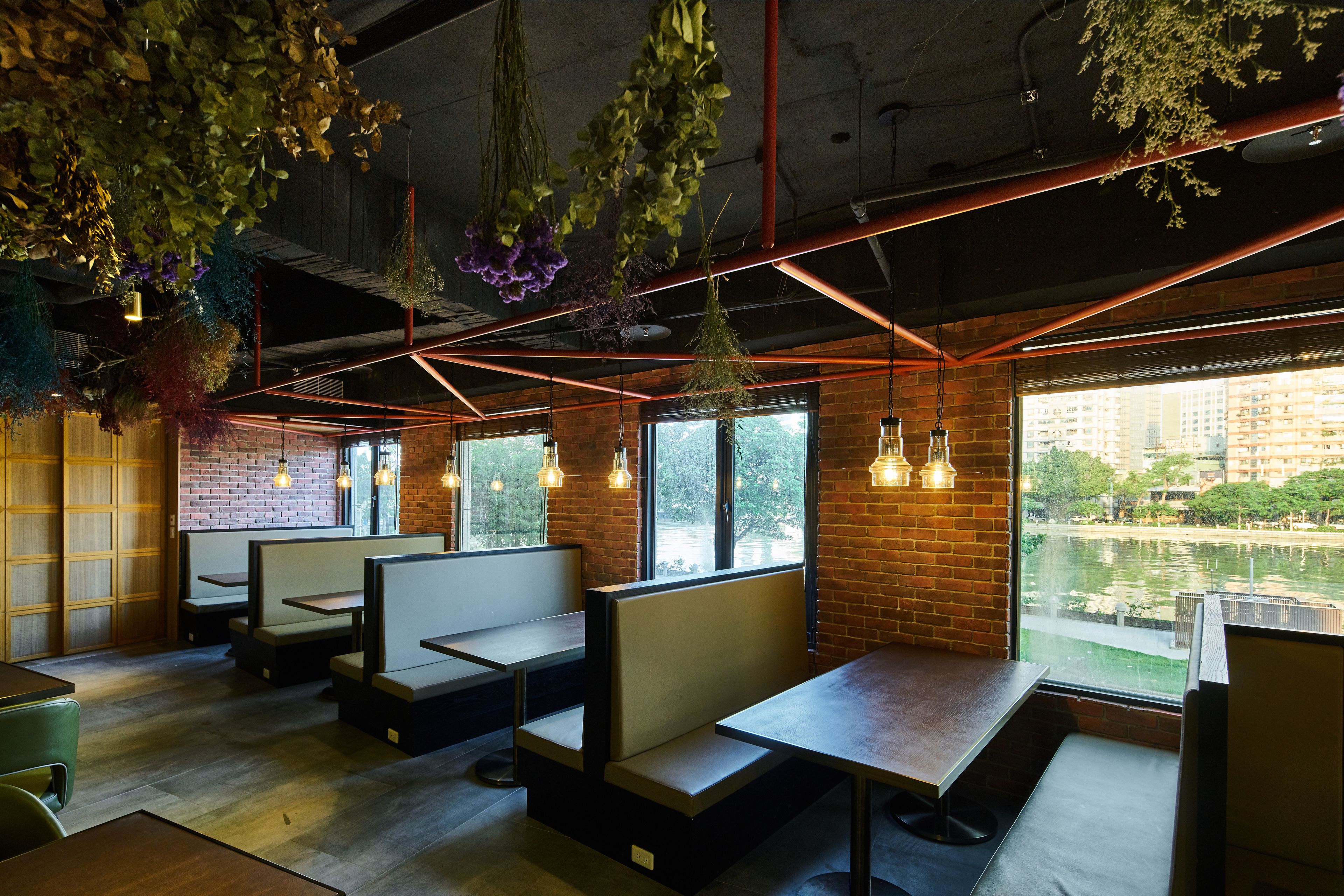 Un restaurant avec des banquettes et des tables, des murs en briques, des plantes suspendues et de grandes fenêtres donnant sur une rivière.