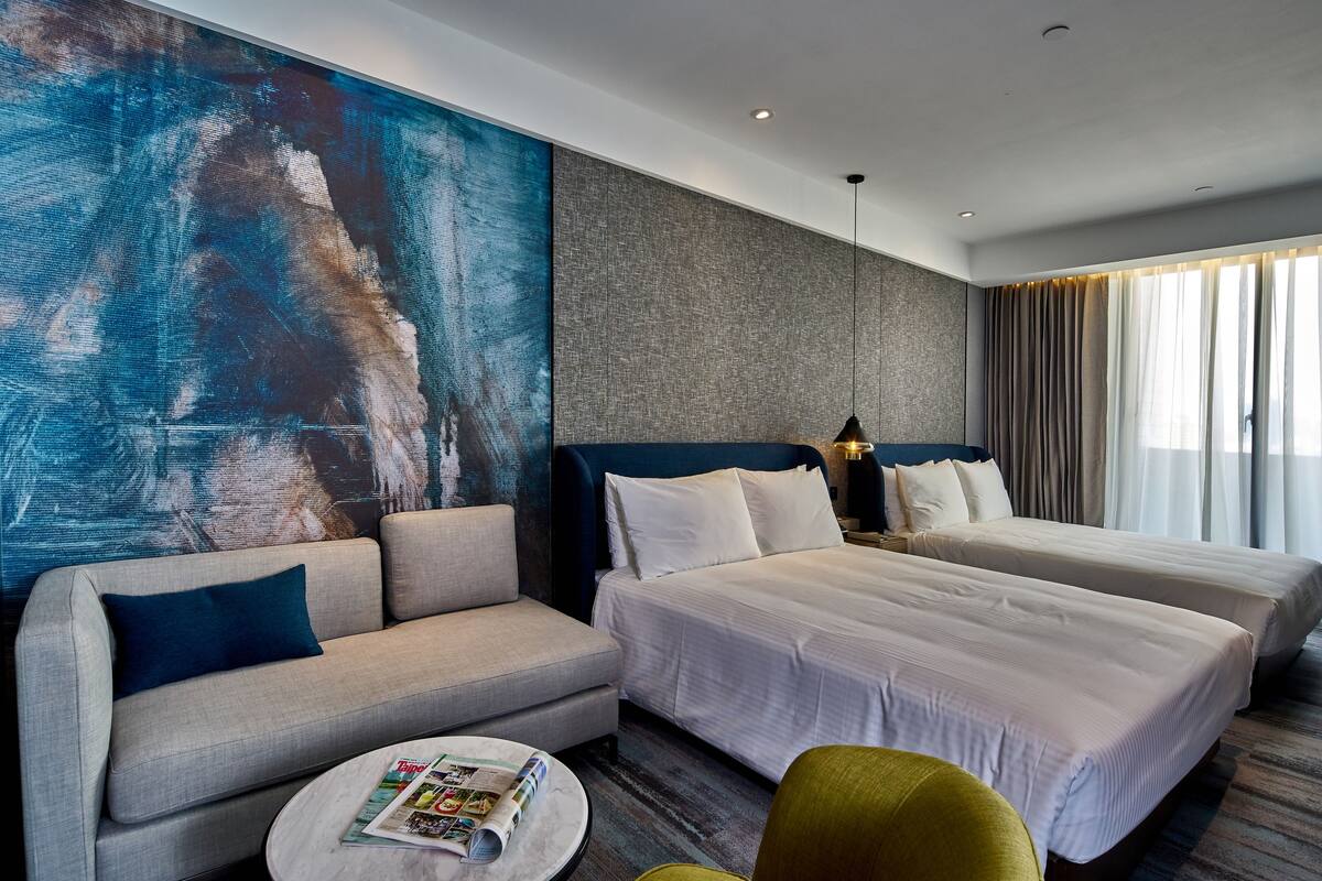 Habitación de hotel con dos camas, un sofá, una mesita de centro redonda y un cuadro abstracto grande en la pared.