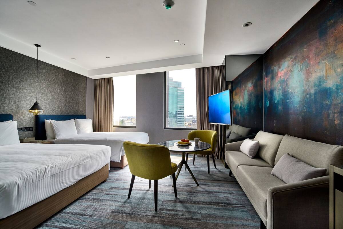 Habitación de hotel con dos camas, un sofá, una mesa de comedor con sillas, un televisor y un amplio ventanal con vista a la ciudad.