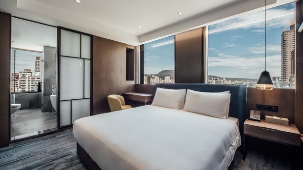 Una habitación de hotel moderna con una cama grande, un escritorio y vistas de la ciudad a través de amplios ventanales.