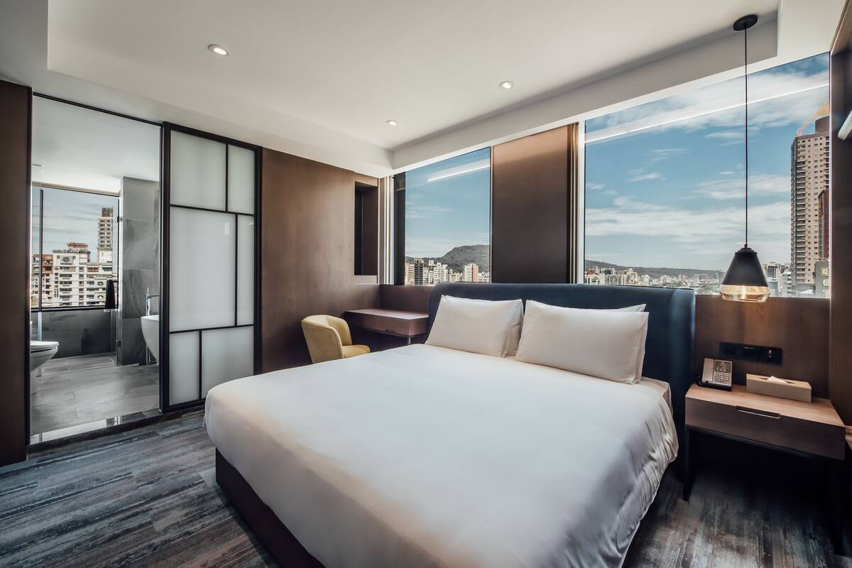 Una habitación de hotel moderna con una cama grande, un escritorio y vistas de la ciudad a través de amplios ventanales.