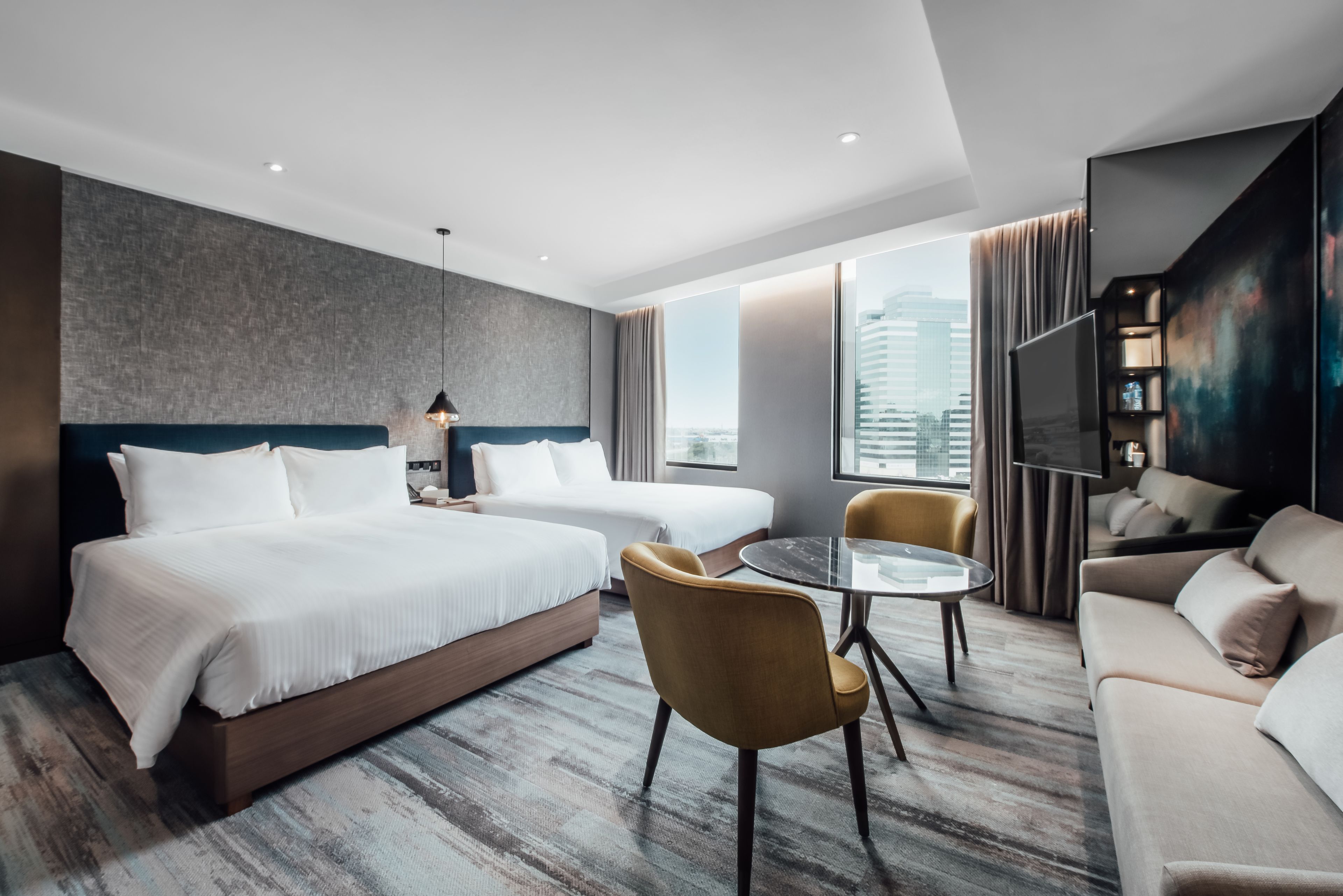 A modern hotel room with a large bed, two chairs, a small table, a TV, and a city view.