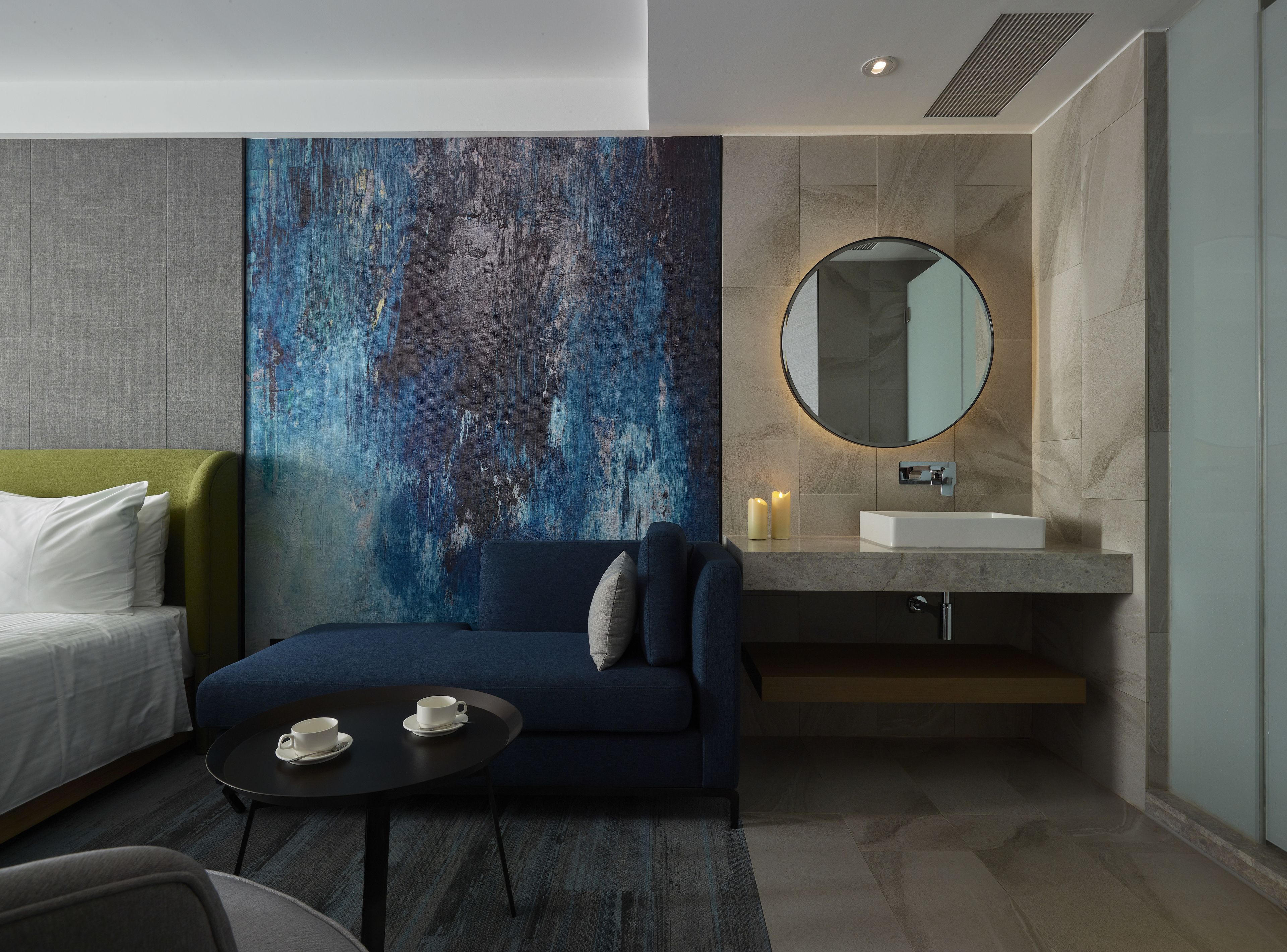 Habitació d'hotel moderna amb un sofà blau, un mirall rodó, una tauleta de nit amb dues tasses i un quadre abstracte gran a la paret.