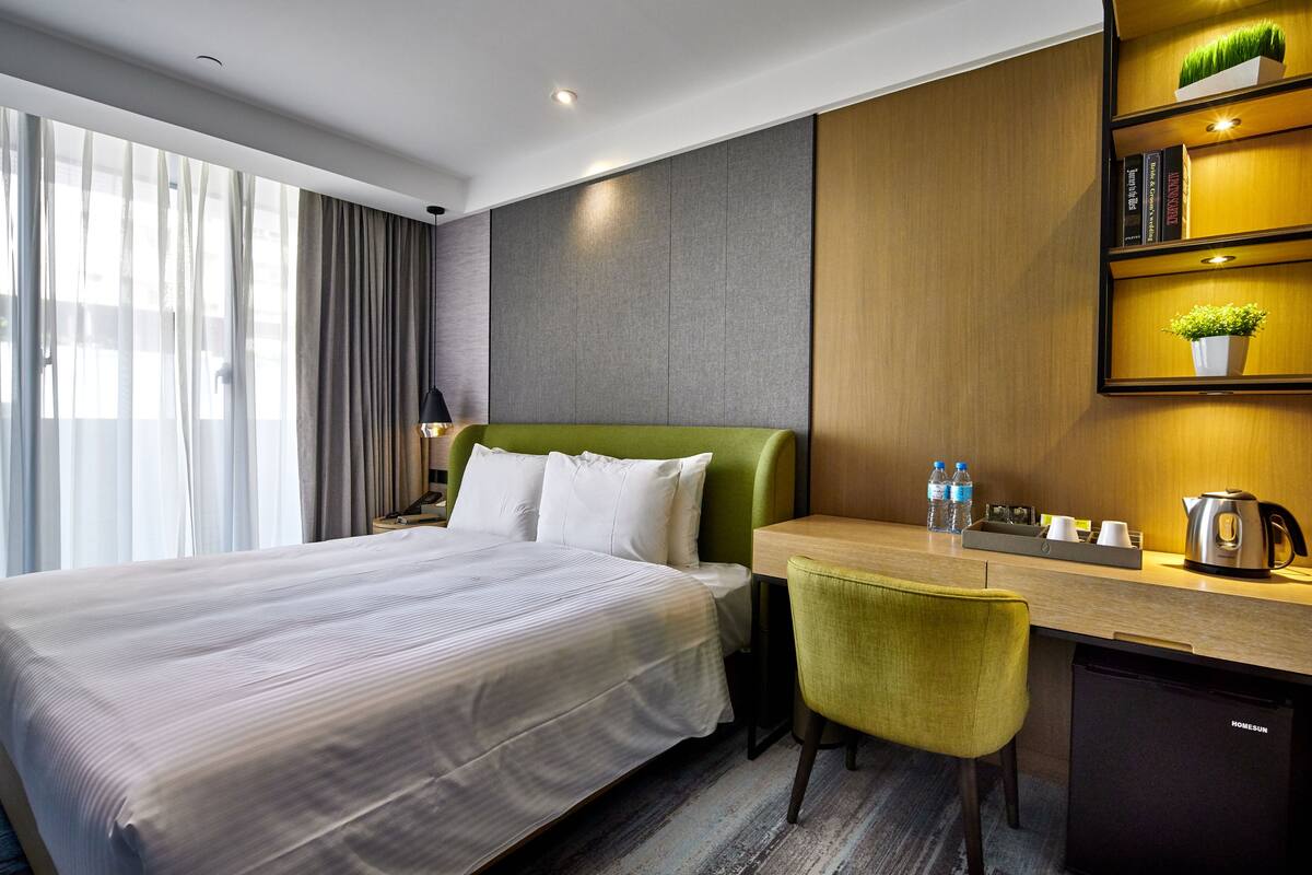 Una habitación de hotel con una cama grande, un escritorio con una silla, una estantería y un pequeño rincón de cocina.