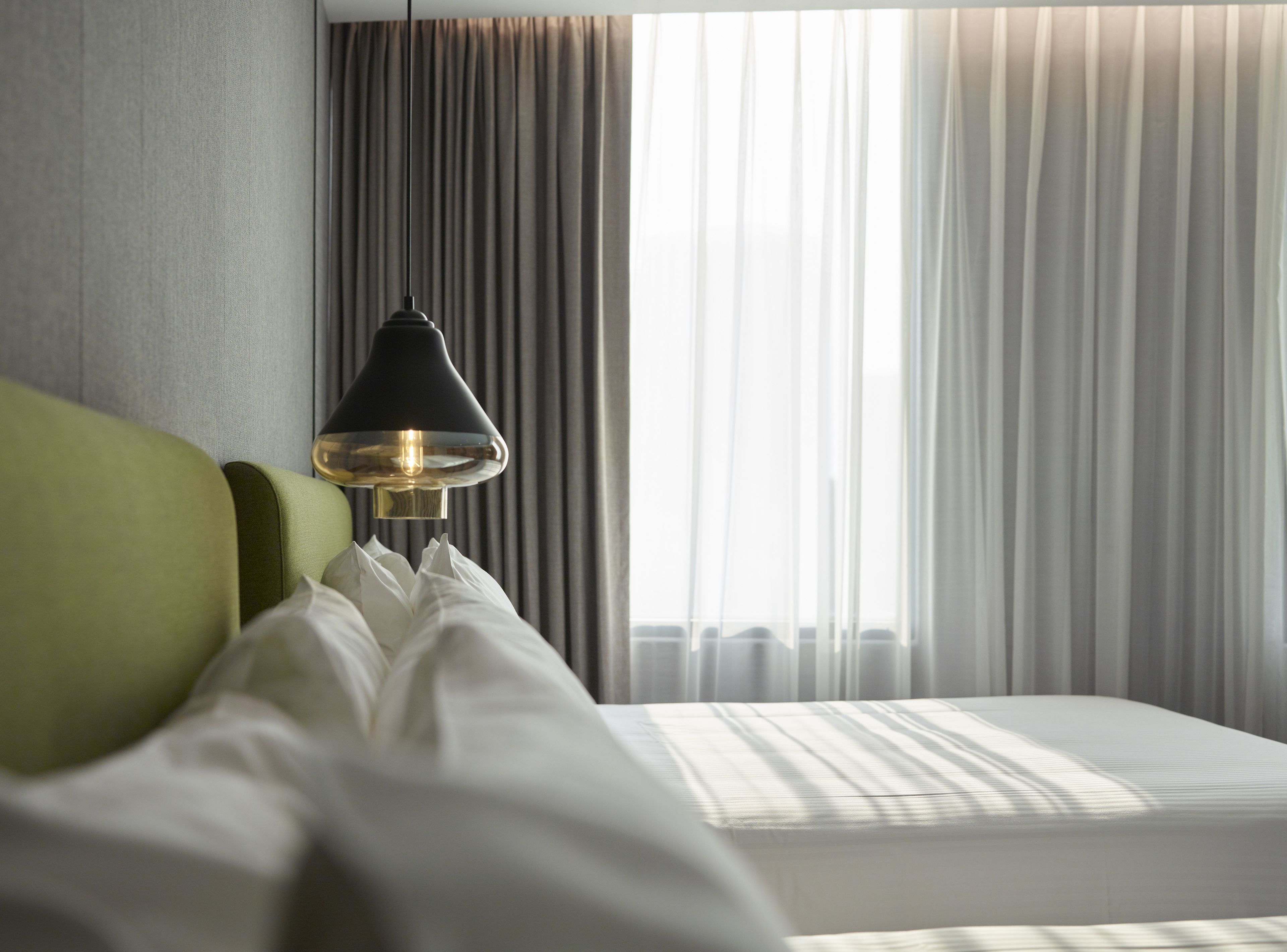 A hotel room with a bed, a green headboard, a black lamp, and sheer curtains.