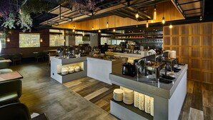 Un restaurante moderno con una zona de buffet central, arreglos de mesas y vistas de la ciudad a través de amplias ventanas.
