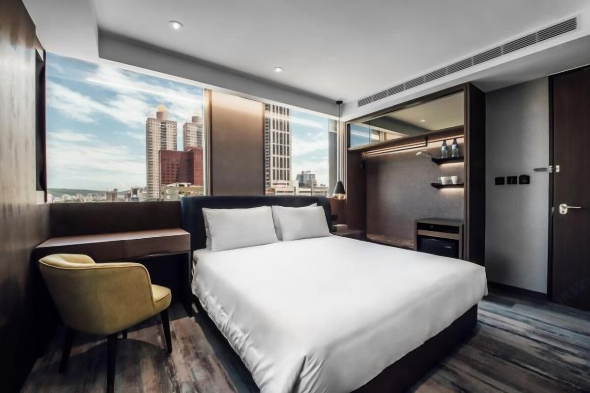 A hotel room with a large bed, a desk, a chair, a wardrobe, and a city view.