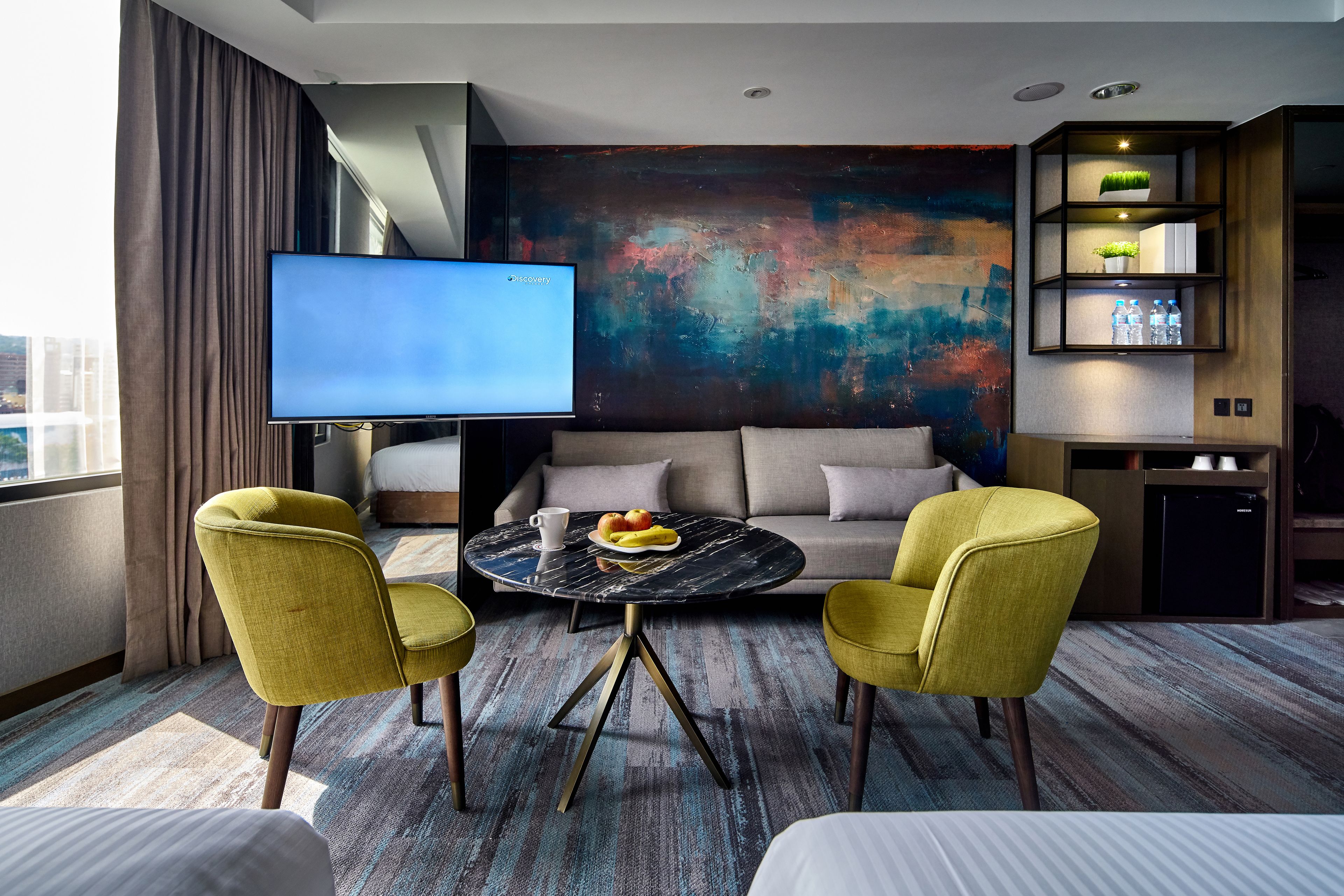 A modern hotel room with a large flat-screen TV, a sofa, two yellow armchairs, a round coffee table with fruit, and a colorful abstract painting on the wall.