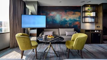 A modern hotel room with a large flat-screen TV, a sofa, two yellow armchairs, a round coffee table with fruit, and a colorful abstract painting on the wall.
