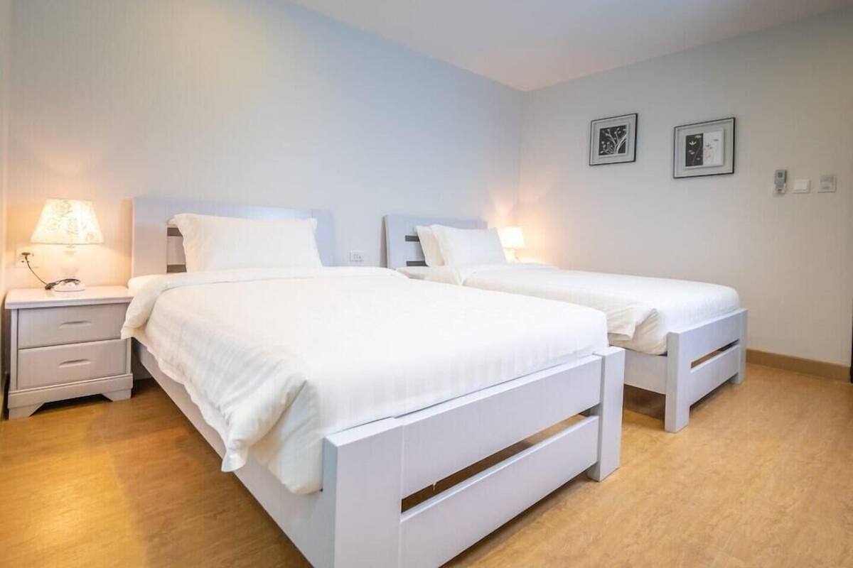 A room with two white beds, a nightstand with a lamp, and a wall with two framed pictures.