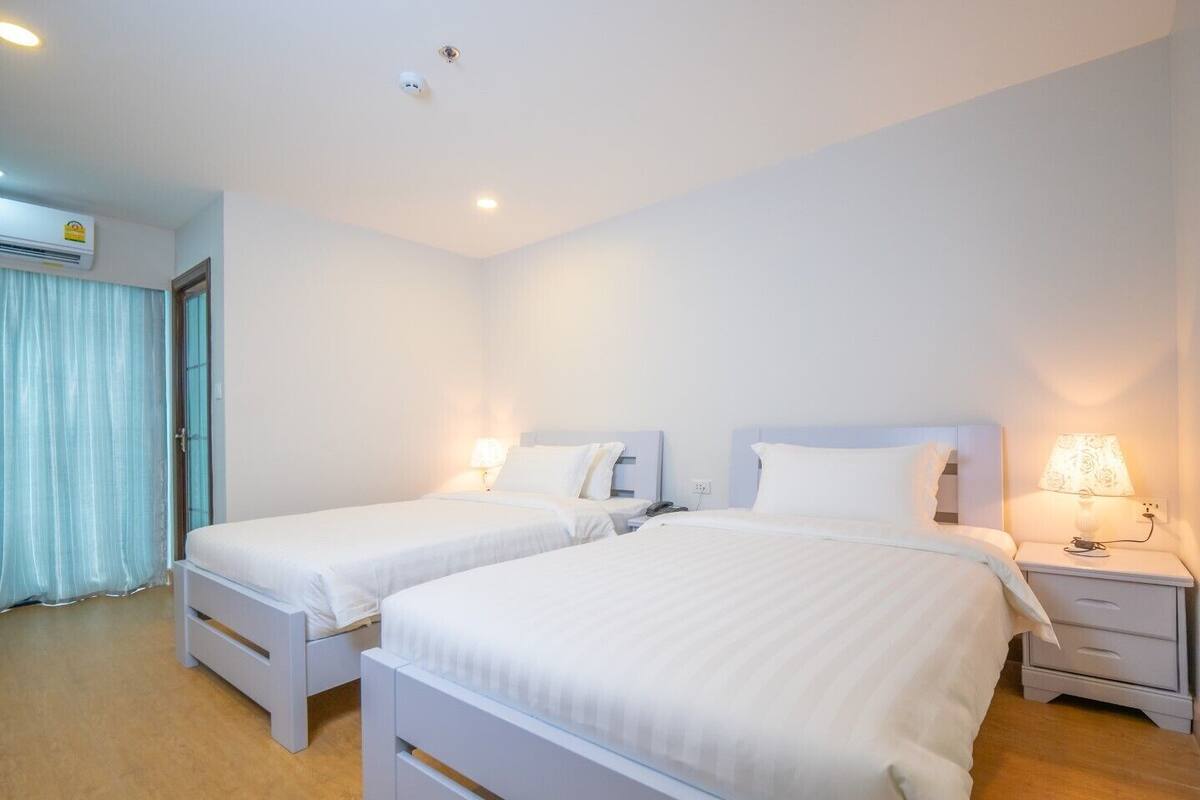 A hotel room with two single beds, a bedside table with a lamp, a window with curtains, and an air conditioning unit on the wall.