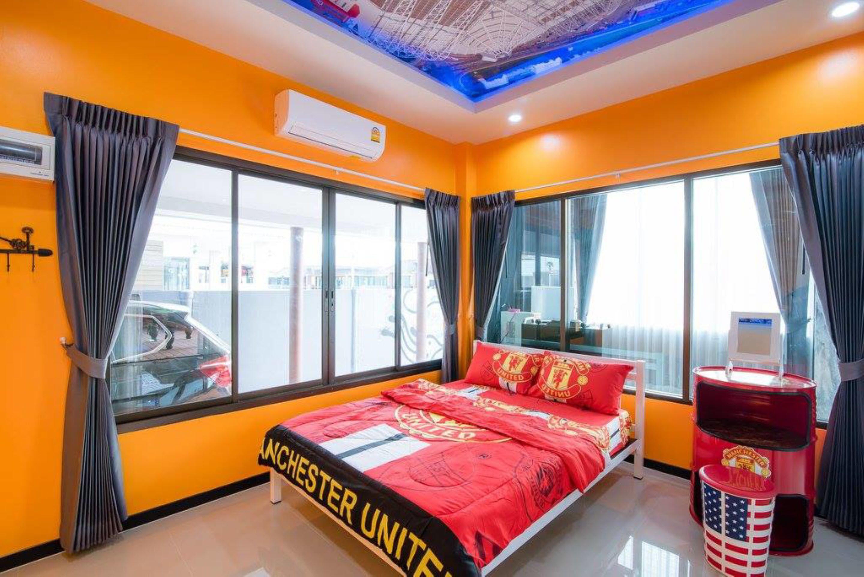 A hotel room with a bed, a red and black Manchester United themed bedspread, a large window, and a ceiling with a blue light design.