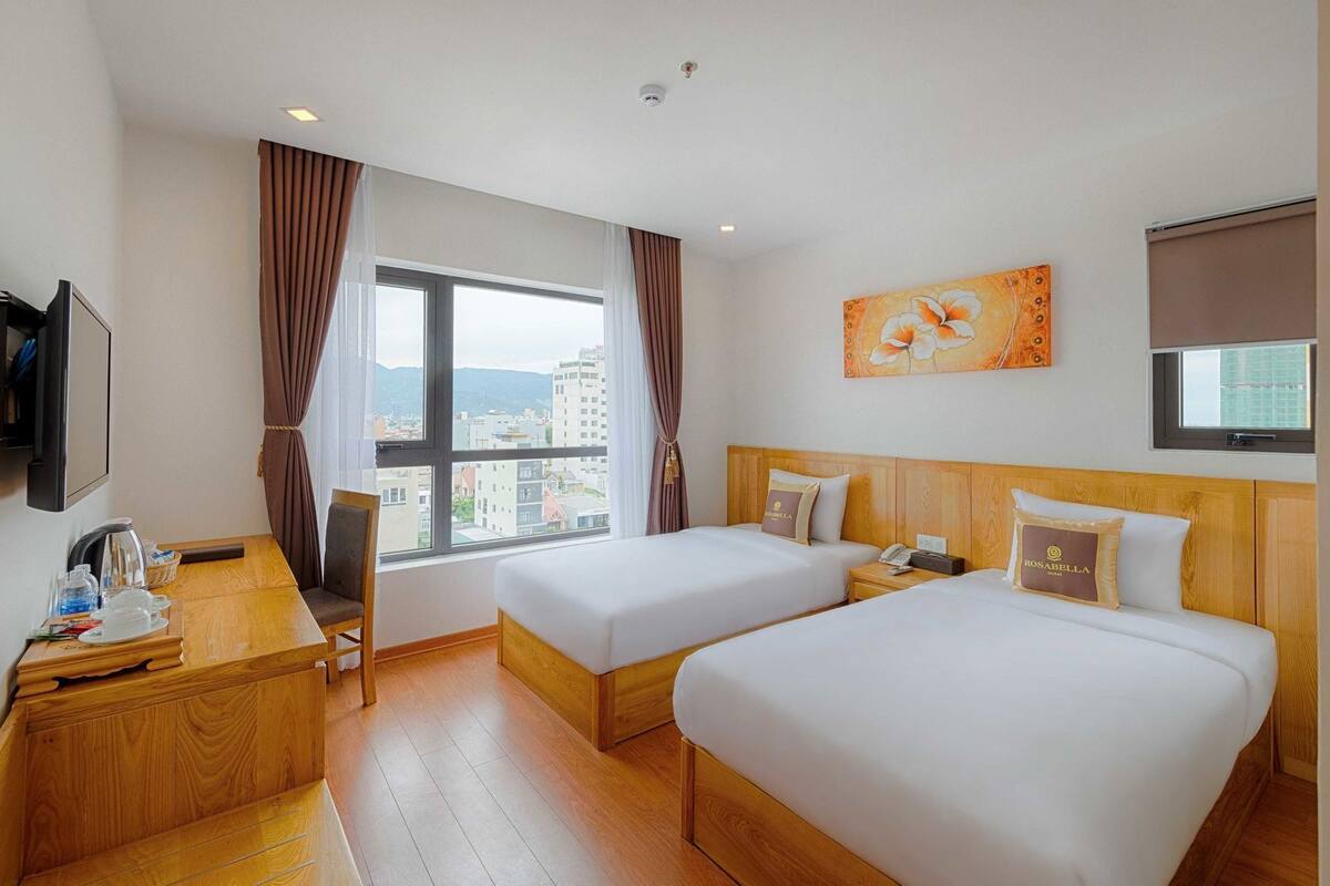 Ein Hotelzimmer mit zwei Betten, einem Schreibtisch, einem Stuhl, einem Fernseher und einem großen Fenster mit Stadtblick.
