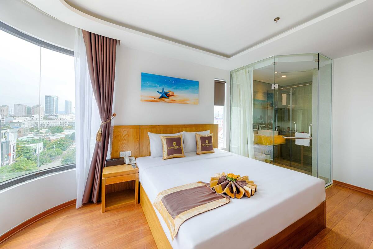 Ein Hotelzimmer mit einem großen Bett, Blick auf die Stadt, einer Duschkabine aus Glas und einem Bild an der Wand.