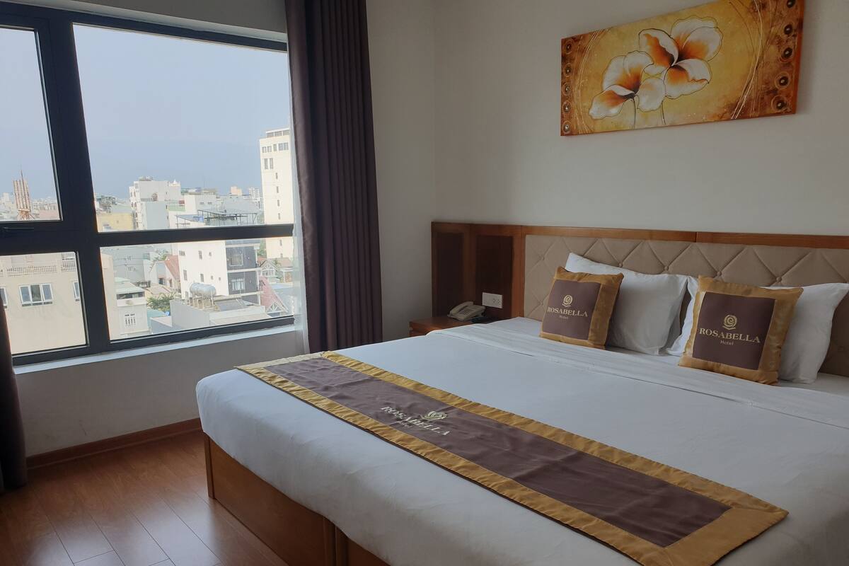 Ein Hotelzimmer mit einem großen Bett, einem Kopfteil, einem Nachttisch, einem Telefon und einem Fenster mit Blick auf die Stadt.