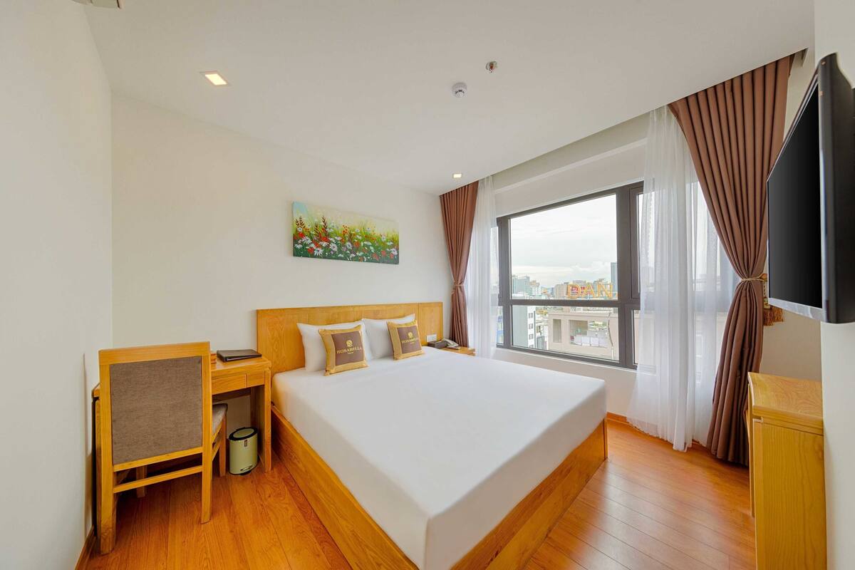 Ein Hotelzimmer mit einem großen Bett, einem Schreibtisch, einem Sessel, einem Fernseher, einem Gemälde an der Wand und einem Fenster mit Vorhängen.