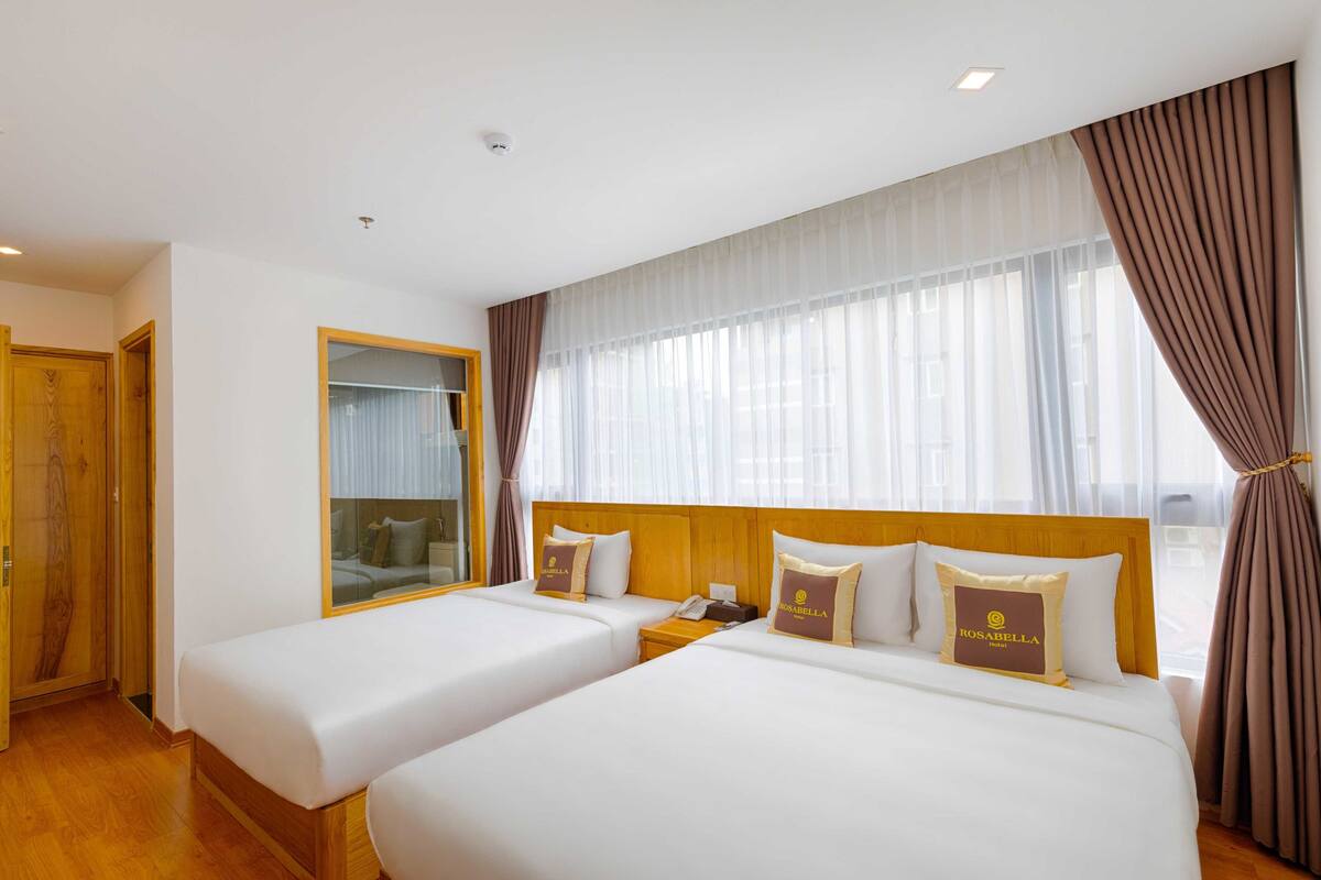 Ein Hotelzimmer mit zwei Betten, einem Holzkopfende, einem Spiegel und einem großen Fenster mit Vorhängen.