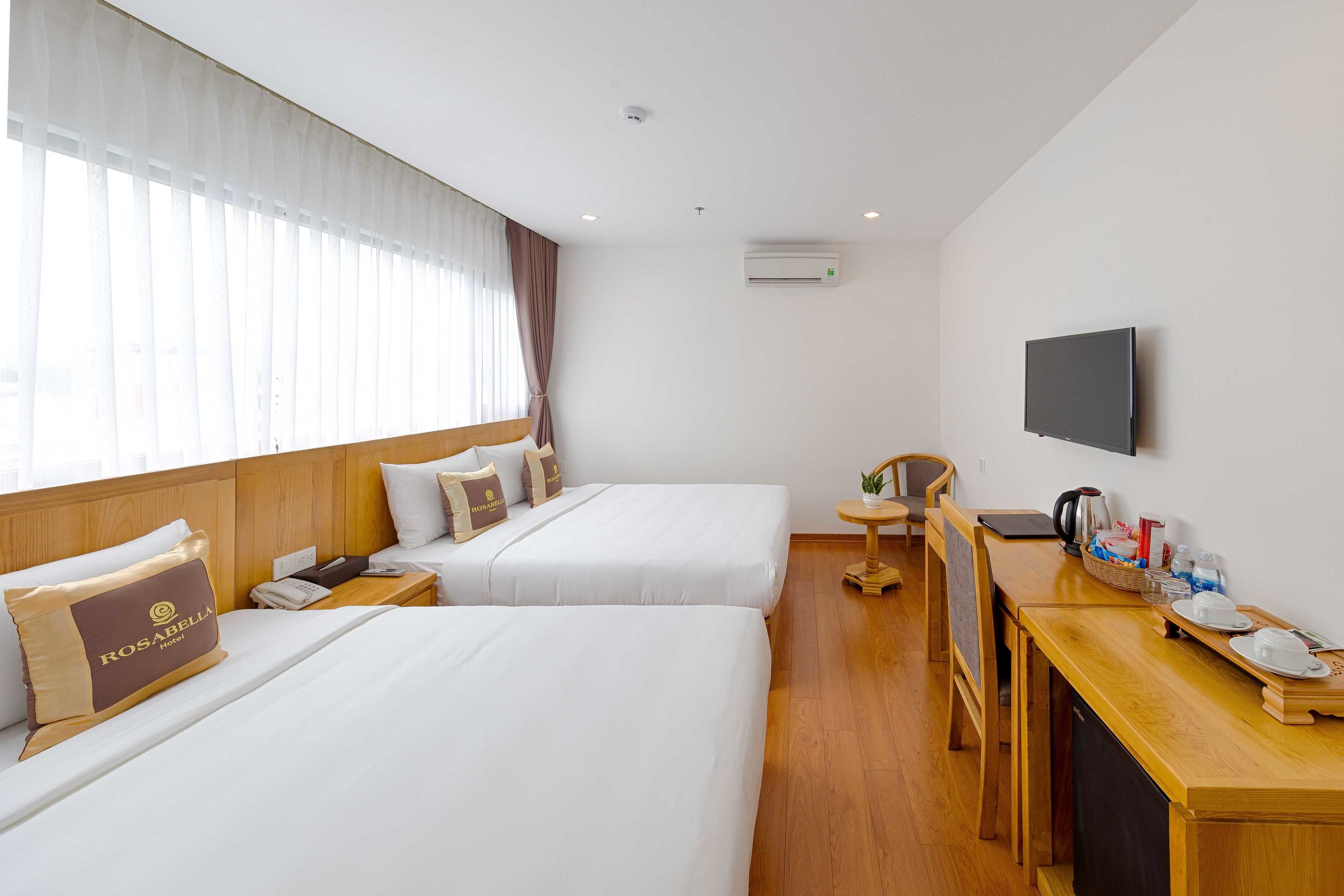 A hotel room with two beds, a wooden desk with a flat-screen TV, and a window with sheer curtains.