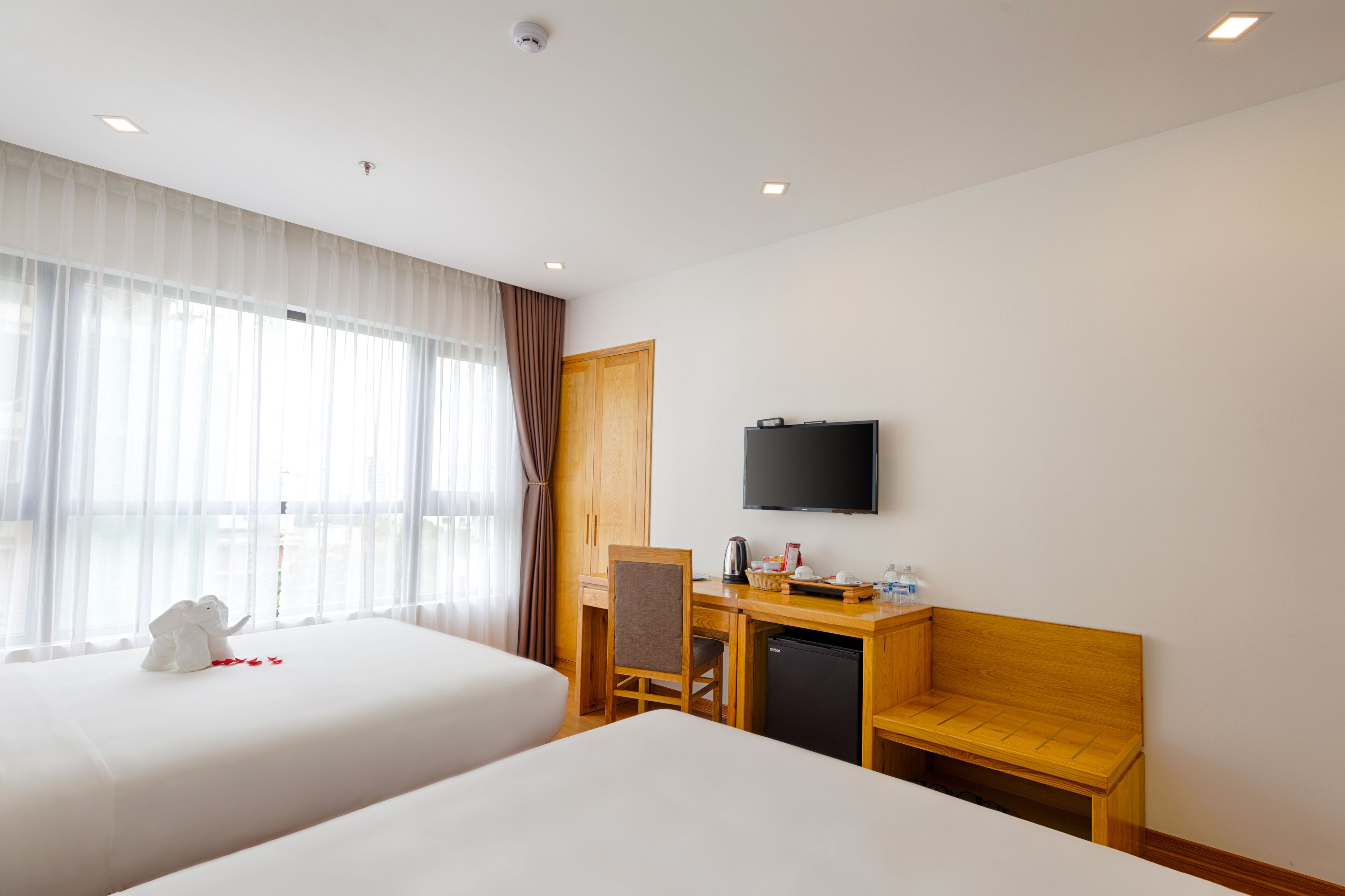 A hotel room with two beds, a desk with a chair, a television, and a small wooden bench.