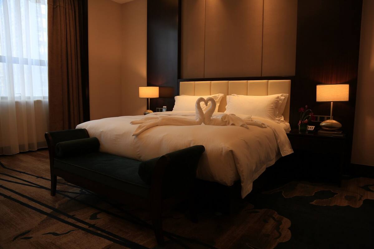 A hotel room with a large bed, two bedside lamps, a nightstand, a chair, and a view of the city through the window.