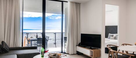 A modern living room with a large window overlooking a balcony, a flat-screen TV, a dining area with wooden chairs, and a sofa.