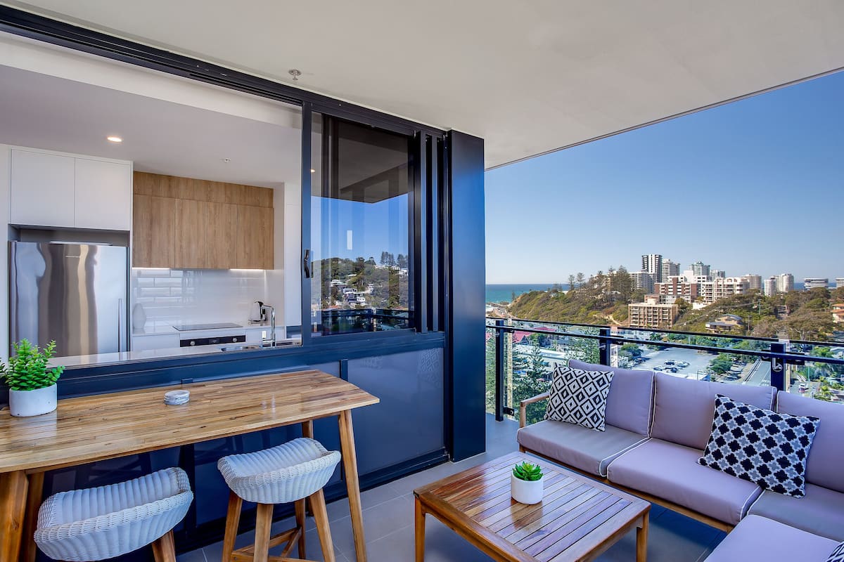 A modern kitchen with a large window, a dining area with a wooden table and chairs, and a balcony with a sofa and cushions.