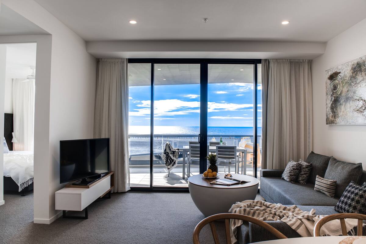 A modern hotel room with a balcony, a flat-screen TV, a sofa, and a dining area.