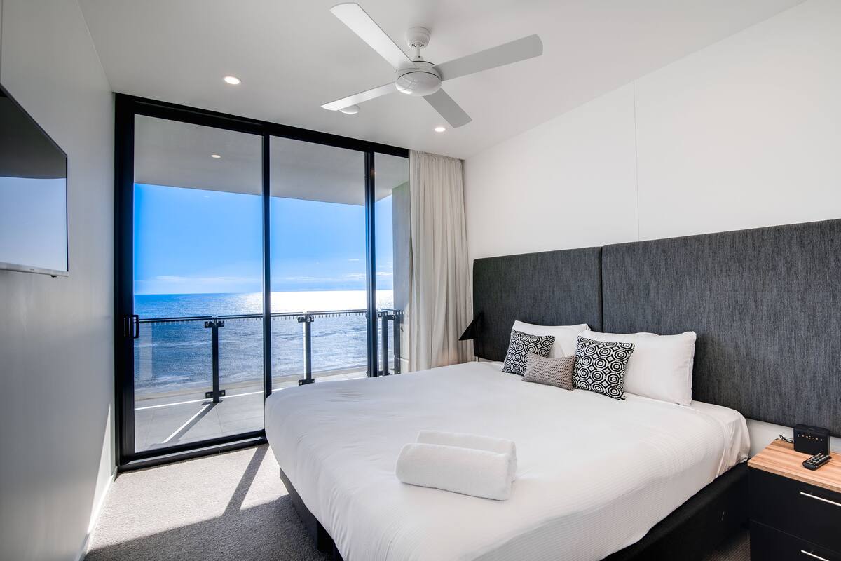 A modern hotel room with a large bed, a ceiling fan, a view of the ocean, and a balcony with a railing.