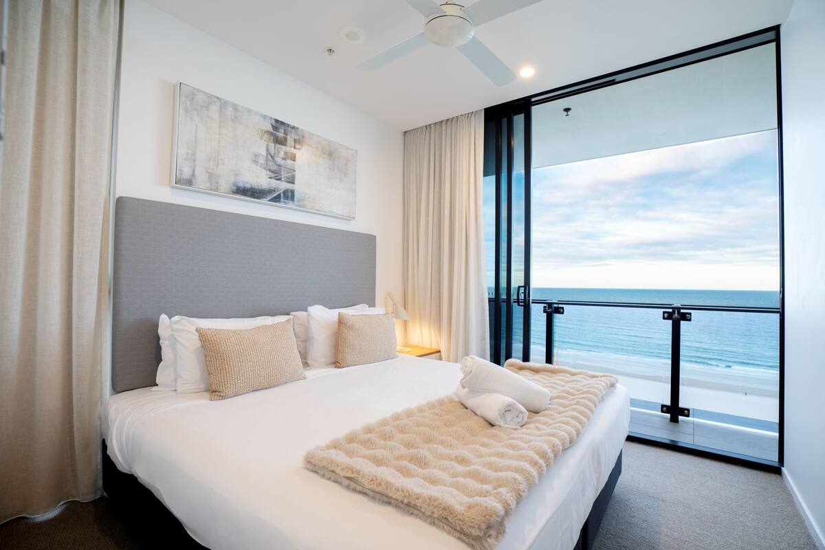 A modern hotel room with a large bed, a ceiling fan, a view of the ocean, and a painting on the wall.