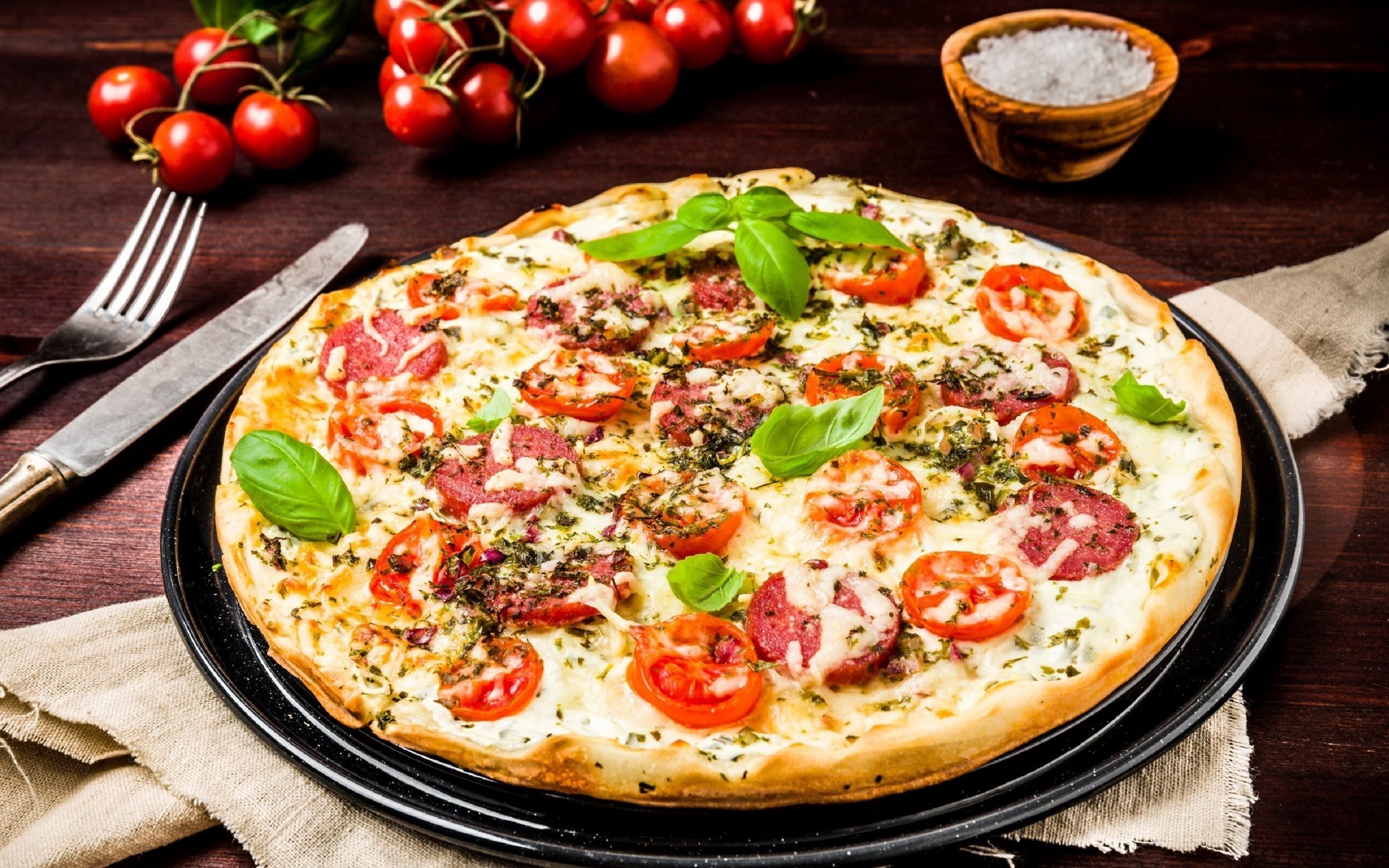 A pizza with various toppings on a black plate, accompanied by a fork and knife, a bowl of salt, and cherry tomatoes in the background.