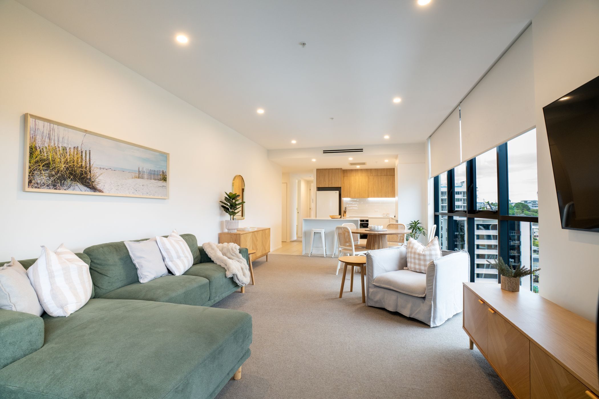 A modern living room with a green sofa, a white armchair, and a wooden cabinet. There is a kitchen area in the background with a dining table and chairs.