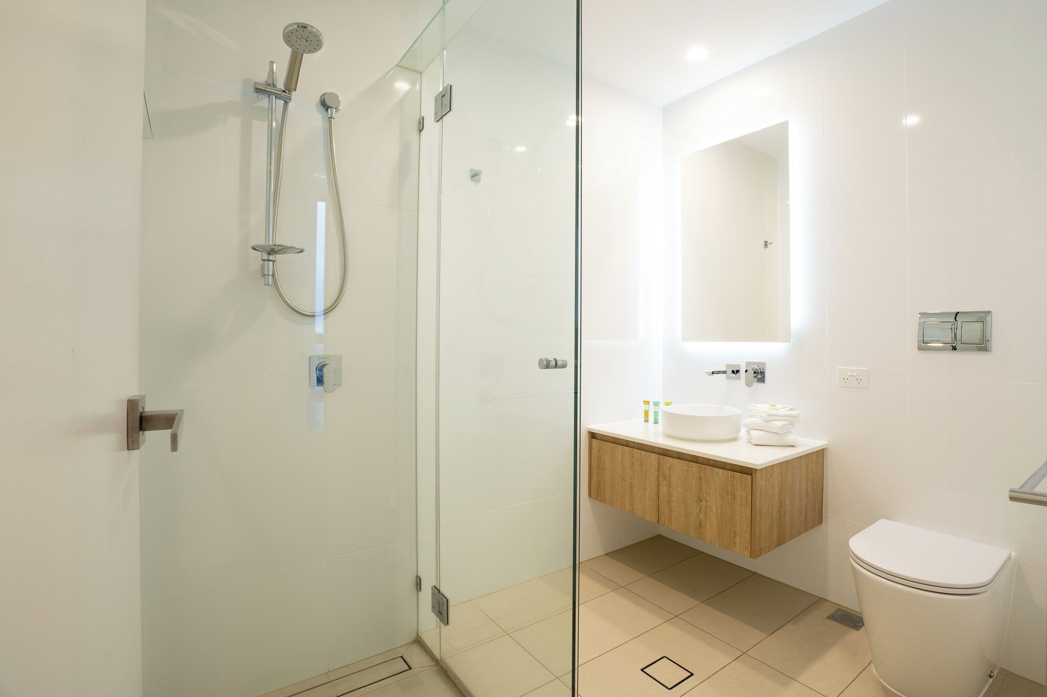 A modern bathroom with a glass-enclosed shower, a wooden vanity, a white toilet, and a mirror above the sink.