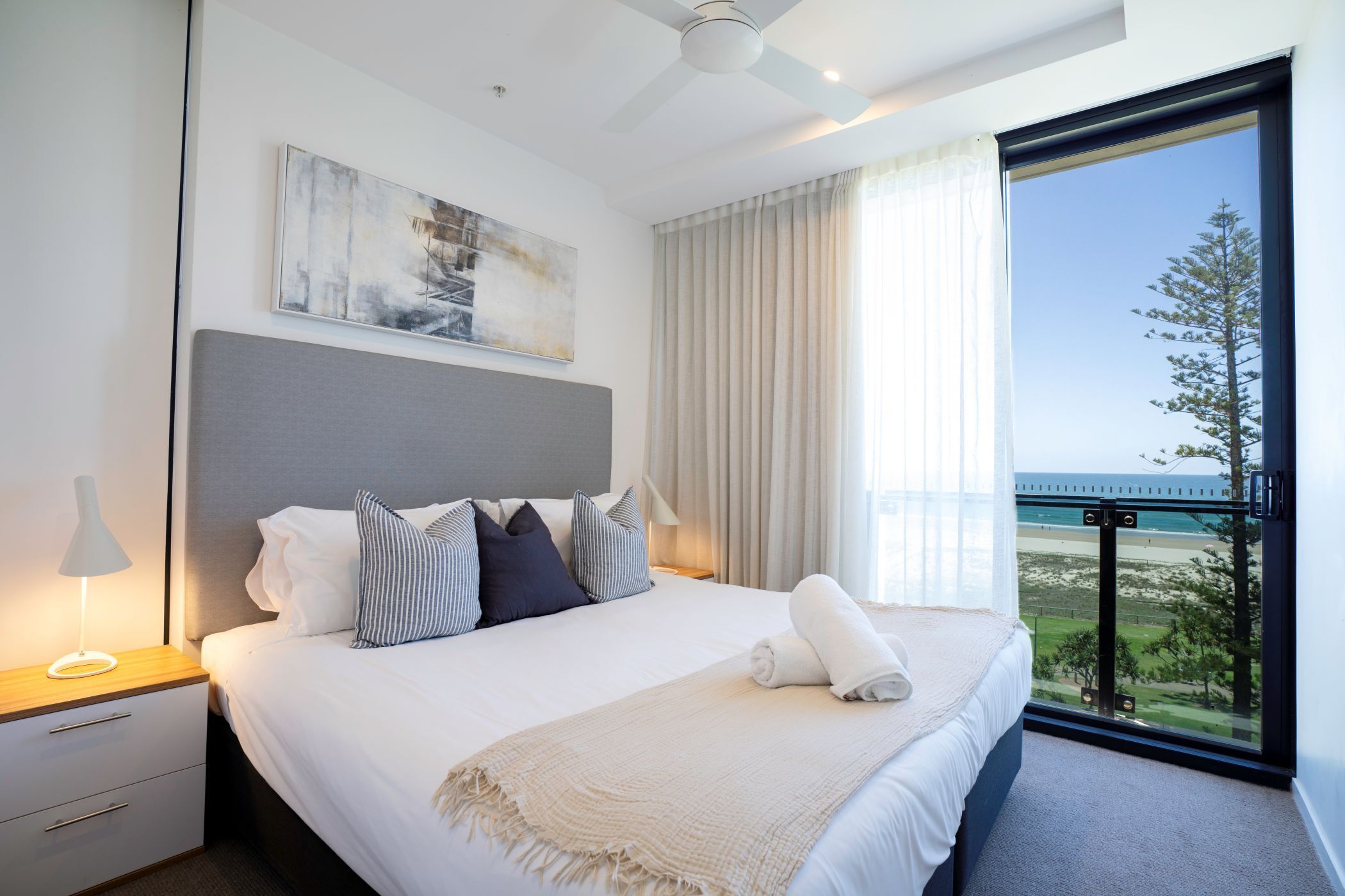 A modern hotel room with a large bed, a nightstand, a lamp, a view of the beach, and a ceiling fan.