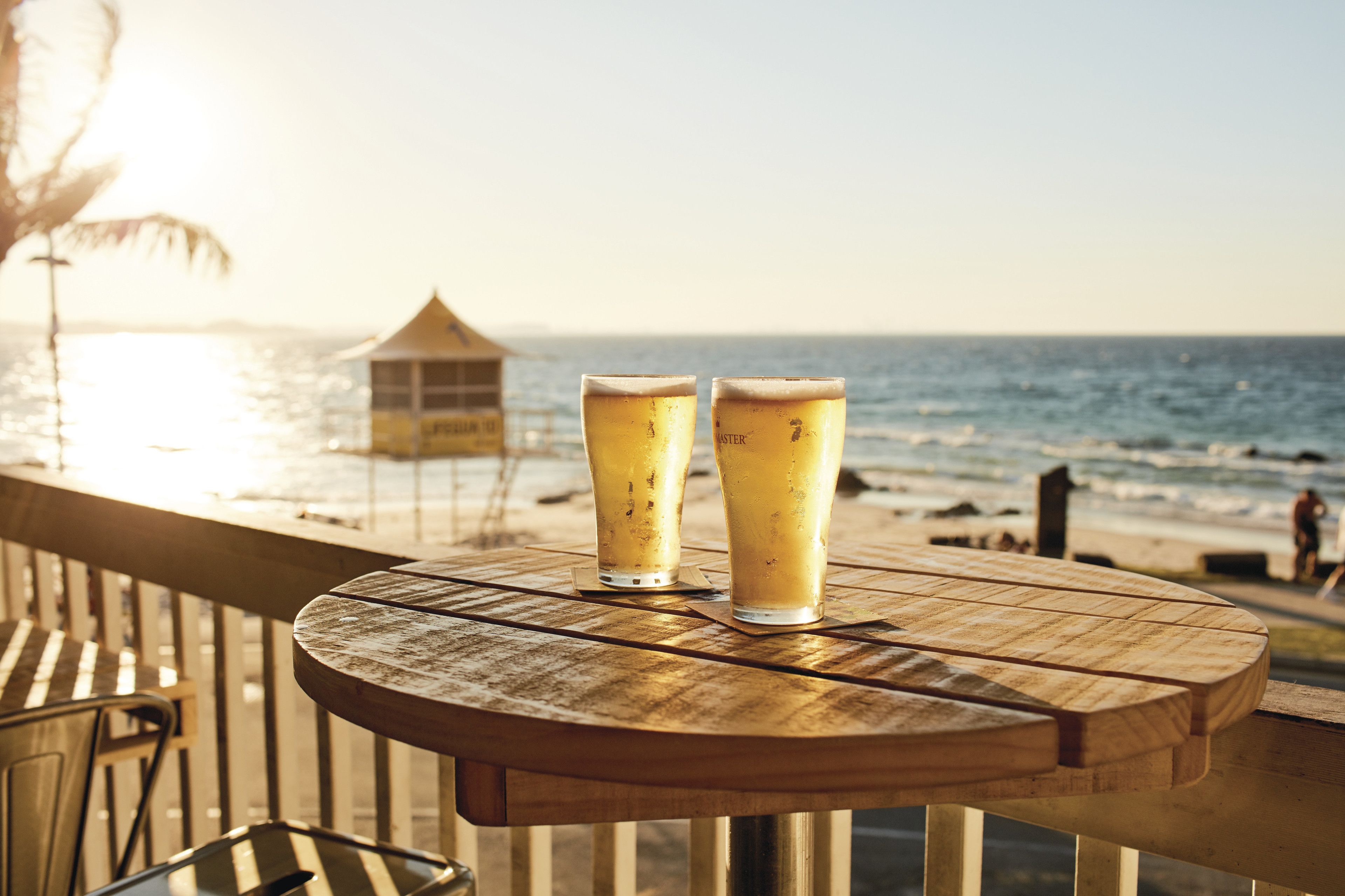 Una mesa de madera con dos vasos de cerveza en un balcón con vista al océano.
