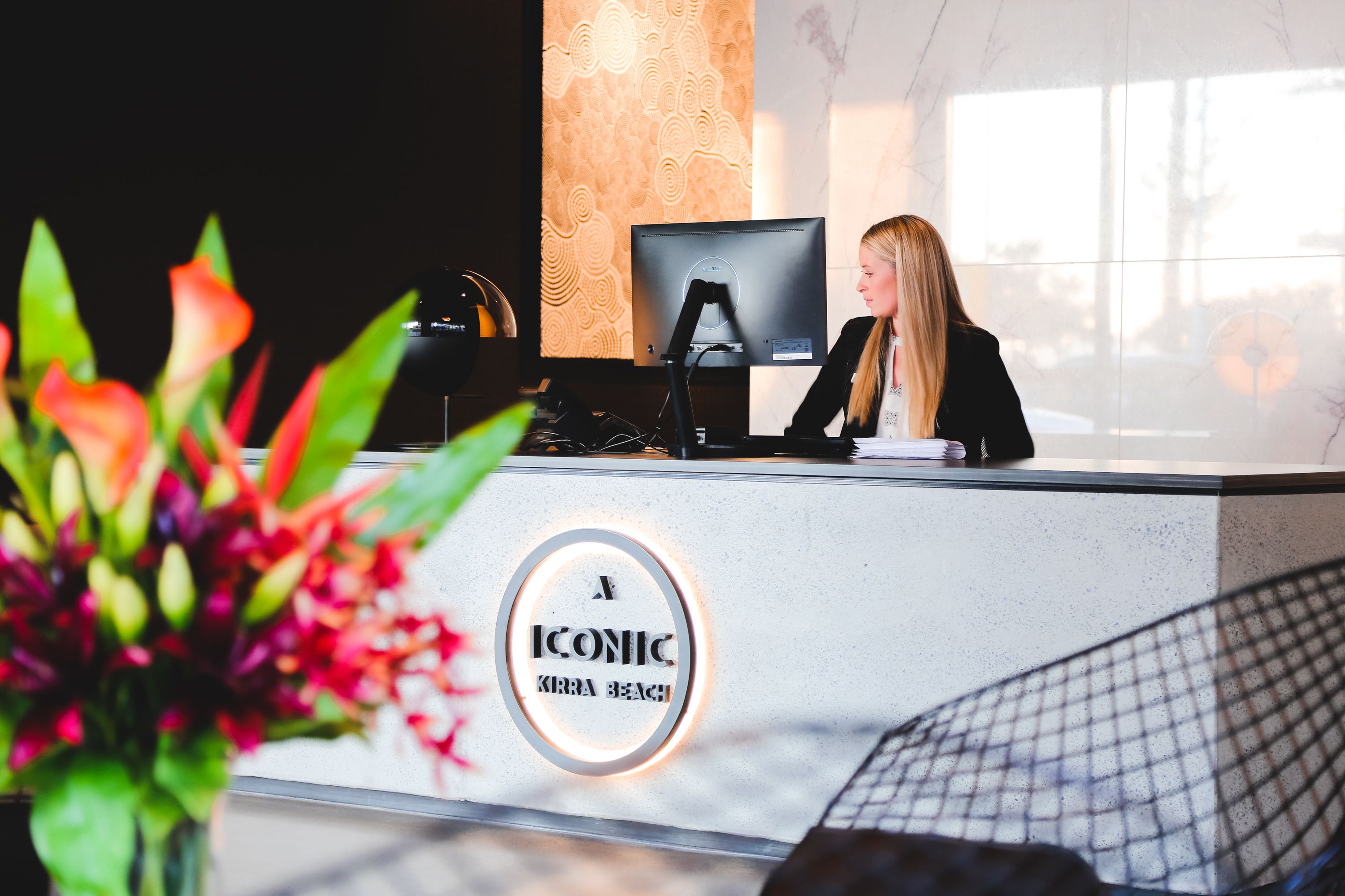 Una mesa de recepción con un monitor de computadora, una mujer sentada detrás y un logotipo que dice "ICONIC KIRRA BEACH".