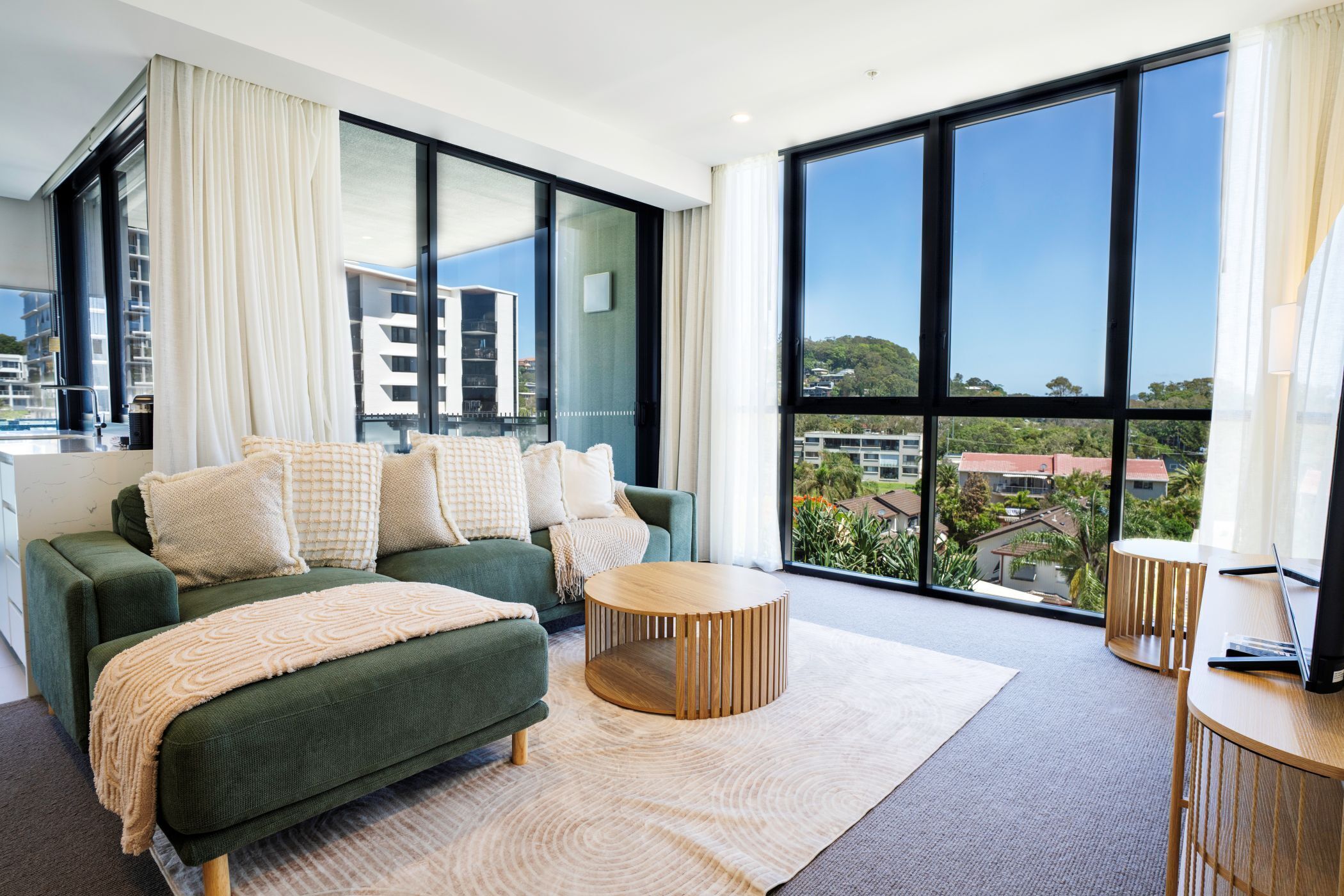 Una sala de estar moderna con un sofá verde, una mesa de centro redonda de madera y ventanas grandes que ofrecen vistas a edificios y vegetación.