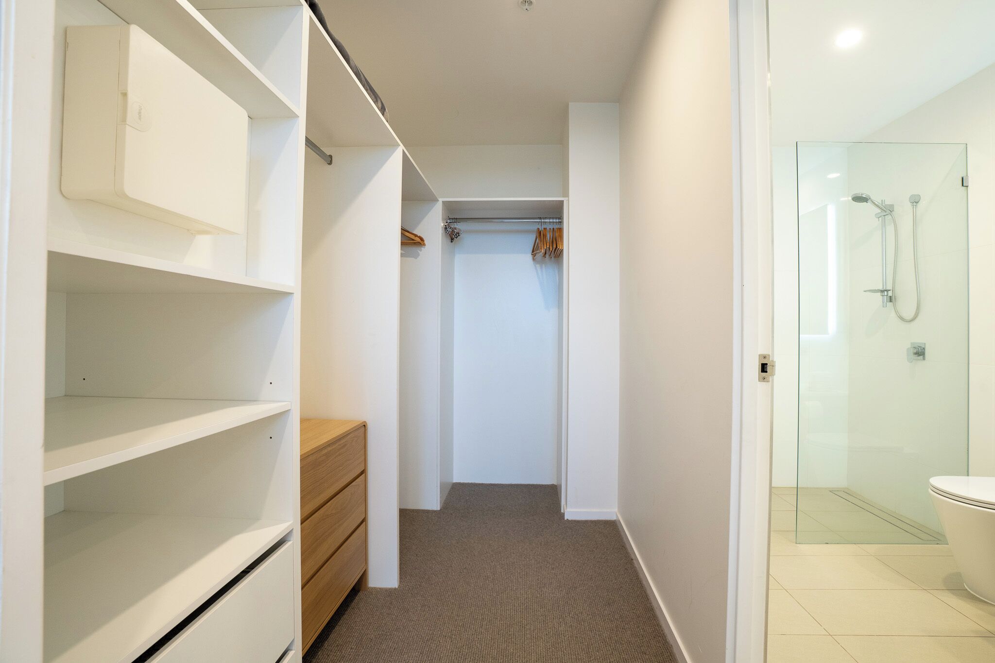 Un baño moderno con cabina de ducha de vidrio, inodoro y un vestidor con estantes y cómoda de madera.