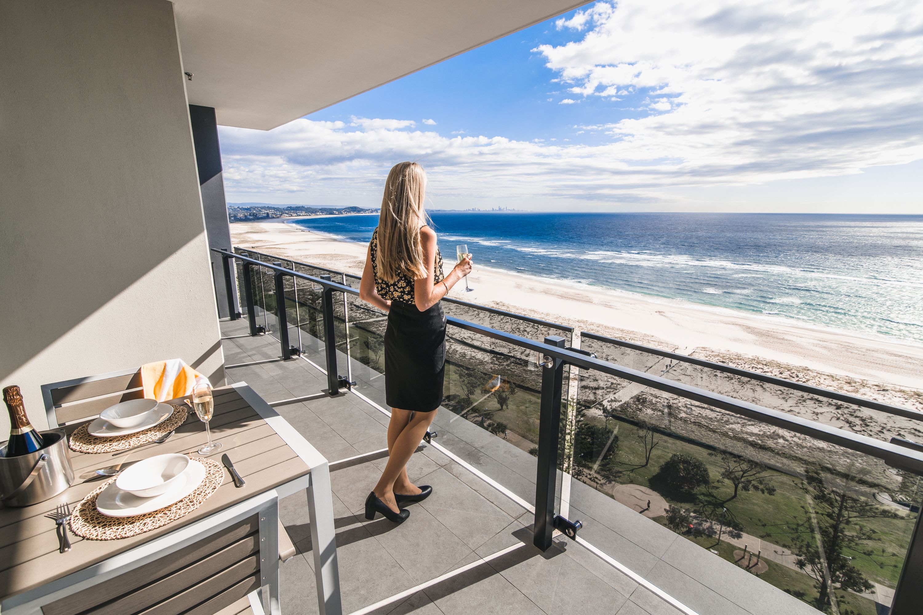 A balcony with a view of the beach, a table set with plates, a bottle of champagne, and a glass.